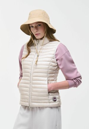 Kobieta w beżowym kapeluszu typu bucket, kremowej pikowanej bezrękawniku i różowej koszuli z długim rękawem, ręce w kieszeniach kurtki, na białym tle.