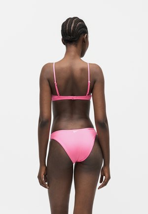 Femme avec des tresses cornrows portant un bikini rose, debout de dos sur un fond clair uni.