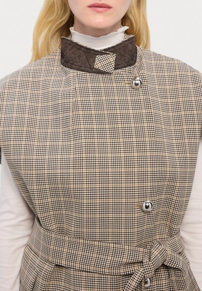 Gilet a scacchi marrone e crema con colletto scuro, dotato di un bottone in argento e dettaglio in vita legato. Tessuto strutturato, scollo alto.