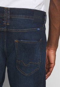 Mørkeblå denimjeans med et tettsittende design, stor baklomme med karakteristisk chevron-søm detalj, og standard beltehalser.