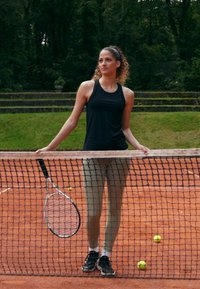 Jogador de ténis em top preto e leggings verde claro apoia-se numa rede com uma raquete de ténis branca. Campo de terra batida laranja com bolas de ténis.