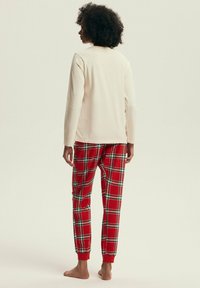 Kremowa bluzka z długim rękawem zestawiona z czerwonymi spodniami dresowymi w kratę, z zielonymi i czarnymi akcentami. Spodnie mają elastyczne mankiety, widoczne z tyłu.