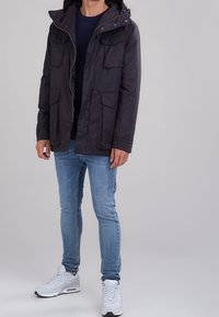 Svart huva jacka med två bröstfickor och två sidofickor, med dragkedja och tryckknappsstängning, bärs över en marinblå topp och ljusblå jeans.