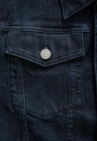 Dark blue denim jacket pocket with silver snap button, featuring a triangular flap and visible stitching details. Textured fabric with subtle sheen.