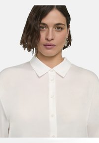 Blusa bianca a maniche lunghe realizzata in tessuto leggero, con chiusura a bottoni sul davanti e colletto classico. Texture morbida con un aspetto trasparente.