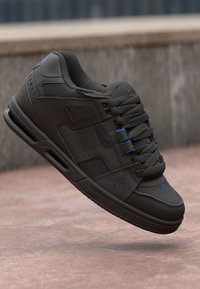 Black sneaker with textured upper, blue accents, flat laces, padded collar, and ridged sole, featuring perforations for breathability.