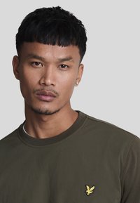 Felpa crewneck verde oliva con colletto a costine, caratterizzata da un logo di un uccello giallo ricamato sul lato sinistro del petto. Tessuto liscio e strutturato.