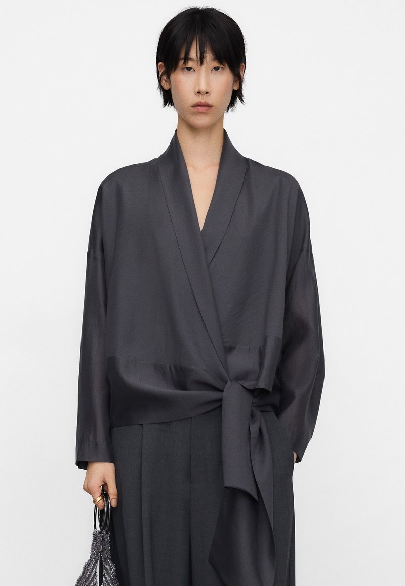 Ciemnoszara bluzka kopertowa o gładkiej satynowej fakturze, z wiązaniem w talii i długimi rękawami, zestawiona z wysokim stanem spodni.