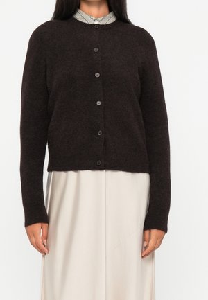 Femme portant un cardigan en laine marron foncé boutonné sur une jupe beige et une chemise rayée avec col.