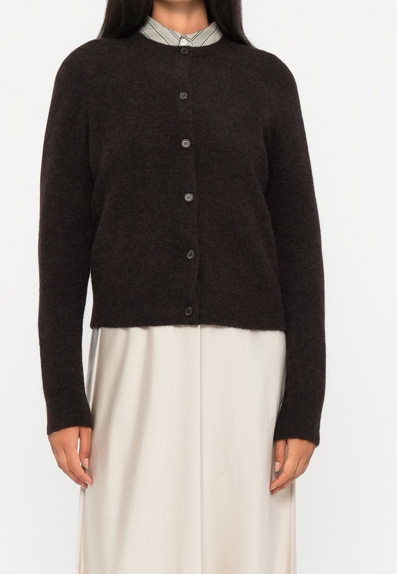 Femme portant un cardigan en laine marron foncé boutonné sur une jupe beige et une chemise rayée avec col.