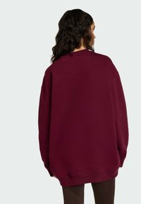 Žena s dlouhými tmavými vlasy, oblečená v oversized vínovém svetru, viděná zezadu na jednobarevném světlém pozadí.