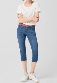 Jeans cortos azules de mezclilla, combinados con una camiseta blanca y un cinturón naranja; llevados con zapatillas blancas. Diseño sencillo, ajuste casual.