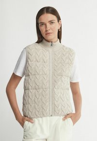 Beige gestrickte Weste mit Zopfmuster, hohem Kragen und Reißverschluss vorne. Getragen über einem weißen Hemd, mit lockerer Passform und seitlichen Taschen.