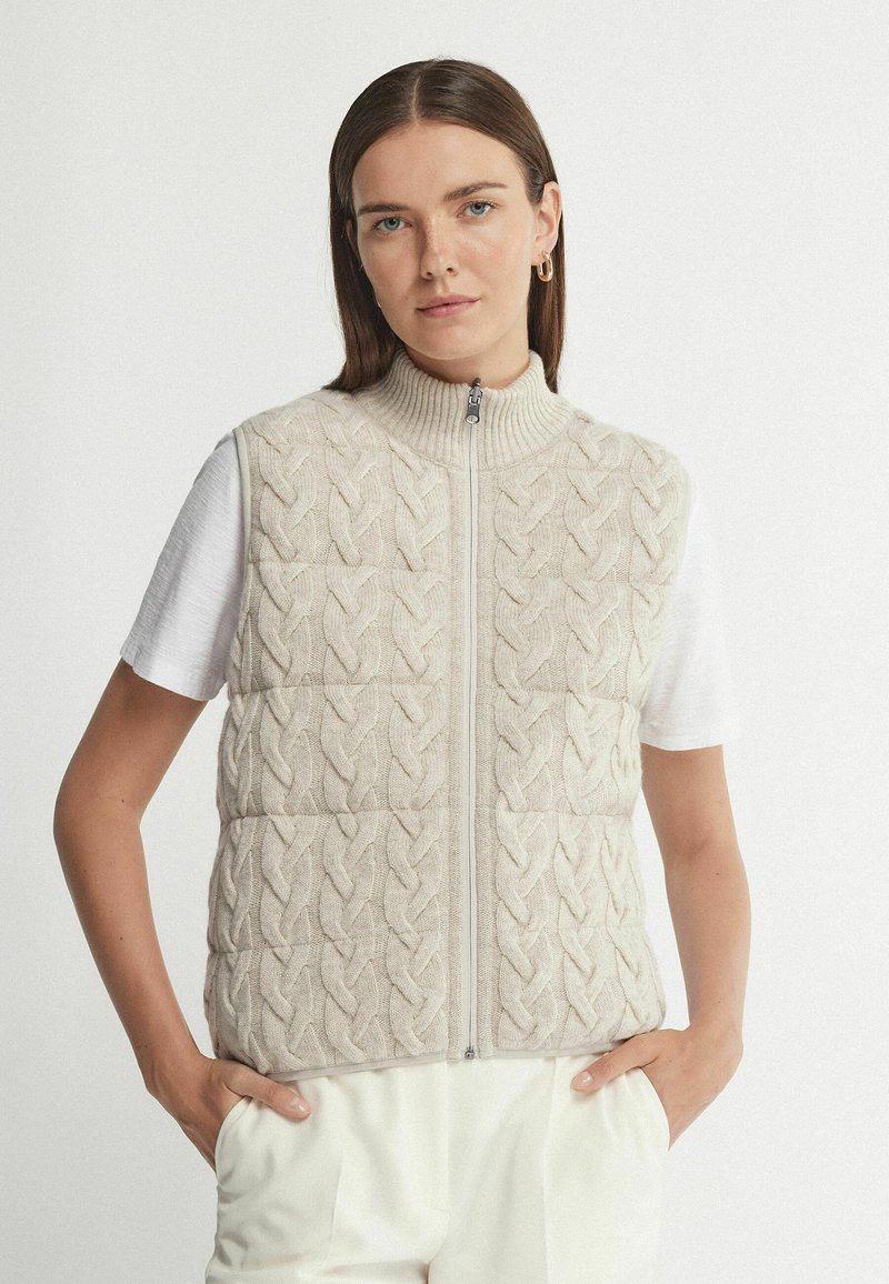 Beige gestrickte Weste mit Zopfmuster, hohem Kragen und Reißverschluss vorne. Getragen über einem weißen Hemd, mit lockerer Passform und seitlichen Taschen.
