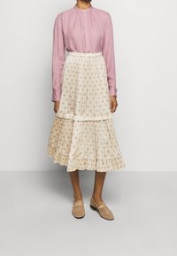 Blusa rosa de manga larga con escote plisado, combinada con una falda crema de flores en capas. El modelo lleva unos zapatos de slip-on marrón claro.