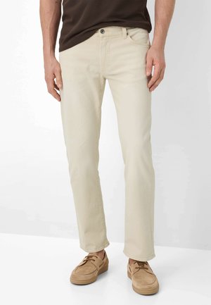 Mann trägt beige, gerade geschnittene Hose, braunes Hemd und beige Schnürschuhe, steht vor einem schlichten weißen Hintergrund.