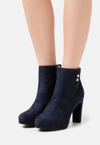 Botas de tobillo de ante azul marino con tacón cuadrado, cremallera lateral y dos botones decorativos dorados cerca del tobillo. Textura suave, diseño ajustado.