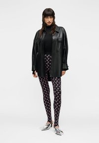 Γυναίκα που φοράει μαύρο, oversized δερμάτινο πουκάμισο, μαύρο τοπ, μαύρο κολάν με ροζ ημισεληνοειδείς και ασημένια μυτερά παπούτσια, με φόντο απλό.