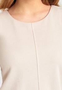 Ljusbeige topp med rund halsringning, texturerad tyg och centralt sömdetalj. Enkel design utan ytterligare accenter eller mönster.