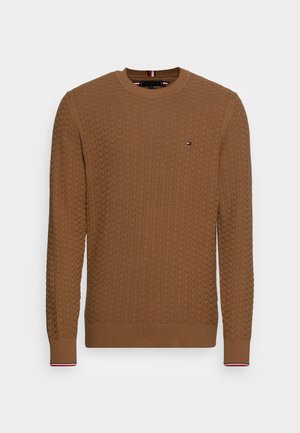 Lichtbruine gebreide trui met textuurpatroon, ronde hals, geribbelde manchetten en klein rood-wit-blauw logo op de borst.