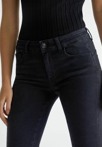 Vaqueros de denim negro ajustados, con una cintura de altura media, bolsillos frontales y cierre de botón metálico. Textura suave sin desgastes visibles.