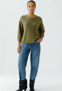 Olive green, loose-fitting knit sweater with three-quarter sleeves, paired with light blue high-waisted jeans and black ankle boots.