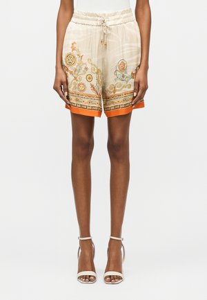 Shorts beige con coulisse, rifiniture arancioni, motivi decorativi di sole e leone, e testo "Just Cavalli" sull'orlo, indossati con sandali bianchi con tacco alto.