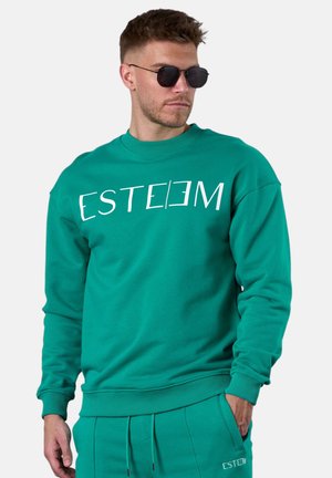 Mann trägt eine türkisfarbene Sweatshirt und passende Hose mit dem Logo "ESTEEM", Hände in den Taschen, schwarze runde Sonnenbrille, schaut zur Seite.