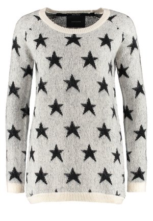 Personne portant un pull gris avec des étoiles noires, un jean skinny foncé, des bottines noires et tenant une sangle de sac à épaule noire.