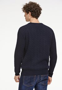 Marineblauer Kabelstrickpullover mit rundem Halsausschnitt, gerippten Bündchen und Saum. Der Stoff wirkt strukturiert; das Design weist vertikale Kabelmuster auf.