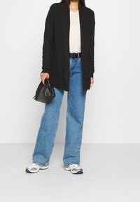 Schwarzer langer Cardigan über einem hellen Top, kombiniert mit lockeren blauen Jeans. Die Accessoires umfassen eine schwarze Tasche und weiße Sneakers mit gelben Akzenten.