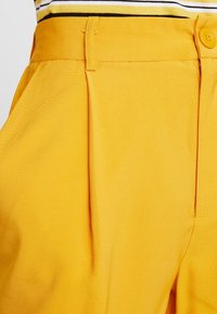 Gula bomullsshorts har skarpa veck, en slät textur och en knäppning i midjan, med synliga sömmar och en krispig finish.