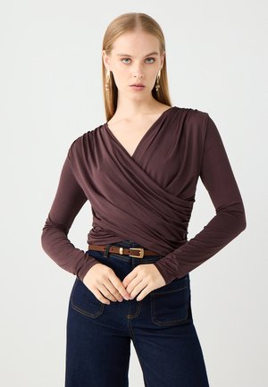Top stile wrap di colore marrone scuro, realizzato in tessuto morbido. Presenta un profondo scollo a V, maniche lunghe e vita aderente. Abbinato a jeans scuri e una cintura.