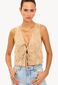 Femme portant un crop top sans manches beige brodé de fleurs noué à l'avant, un jean taille haute bleu clair, des bijoux en or, les mains dans les poches.