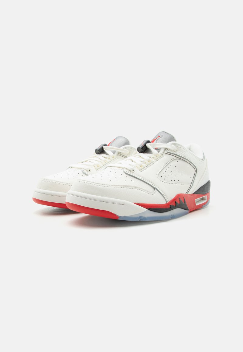 Par de zapatillas blancas de corte bajo con detalles en rojo y negro en la suela, lados perforados y cierres negros en los cordones sobre un fondo gris.