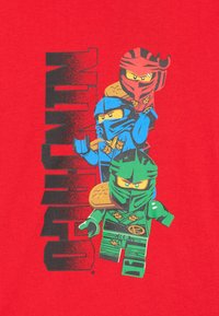 Rotes T-Shirt mit drei cartoonhaften Ninja-Figuren in Blau, Grün und Rot, mit dem grafischen Text "NINJA" in Schwarz auf der Vorderseite.