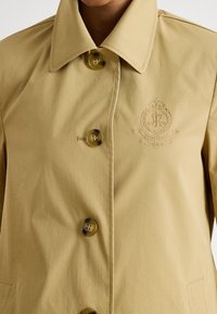 Béžový kabát na zapínanie na gombíky z odolnej tkaniny, s veľkým golierom, jemnou výšivkou a okrúhlymi gombíkmi s motívom. Jednoduchý dizajn s vreckami.