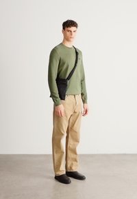 Zöld hosszú ujjú ing, bézs nadrág, fekete cipő és fekete oldaltáska. Egyszerű, texturált ingből és bő szabású nadrágból álló kialakítás.