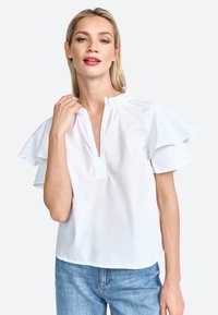 Blusa bianca a maniche corte con dettagli arricciati, scollatura a V e vestibilità rilassata, abbinata a jeans in denim azzurro chiaro.