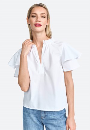 Blouse blanche à manches courtes avec des accents volants, un décolleté en V et une coupe décontractée, associée à un jean en denim bleu clair.
