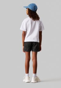 Bambino che indossa una t-shirt bianca in cotone, pantaloni corti di denim neri, calzini bianchi e sneakers beige. Un cappellino azzurro completa l'outfit.