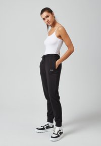 Zwarte sweatpants met elastische tailleband en trekkoord, gecombineerd met een wit tanktop en zwarte sneakers met witte zolen en accenten.