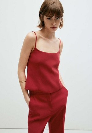 Jeune femme aux cheveux bruns courts, portant un haut rouge sans manches et un pantalon rouge assorti, debout les mains dans les poches sur un fond uni.