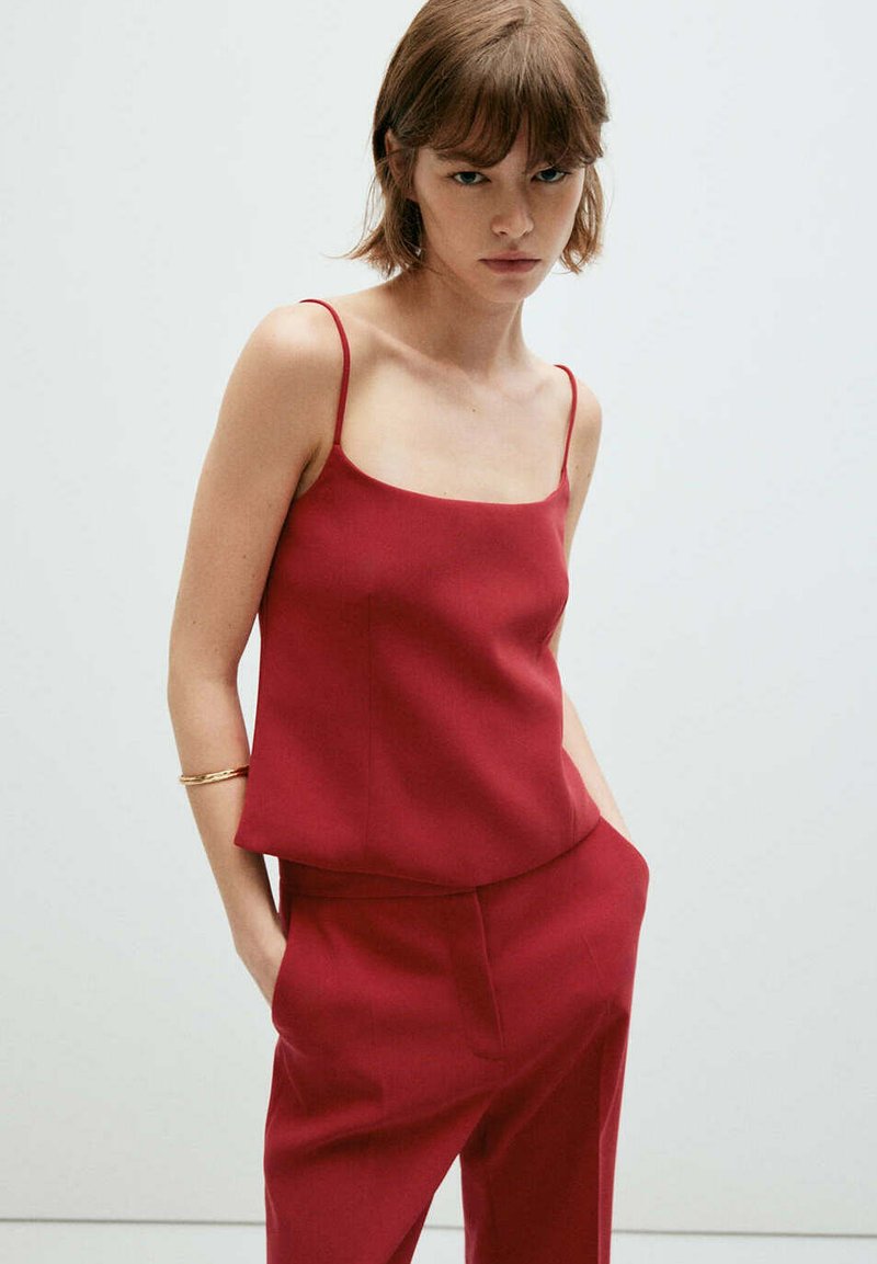 Jeune femme aux cheveux bruns courts, portant un haut rouge sans manches et un pantalon rouge assorti, debout les mains dans les poches sur un fond uni.