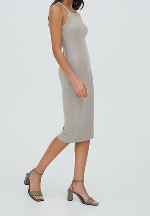 Femme portant une robe midi beige ajustée sans manches et des sandales vertes à talons blocs marchant sur un fond clair uni.