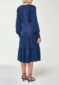 Kvinna iklädd en marinblå midiklänning med lång ärm, spetspaneler och lagerkjol, matchad med svarta, vadderade sandaletter med låg klack, visad bakifrån.