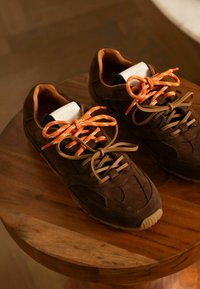 Bruine suède sneakers met oranje en beige veters, ronde neus, gestructureerde bovenkant en gevoerde binnenkant, neergelegd op een houten oppervlak.