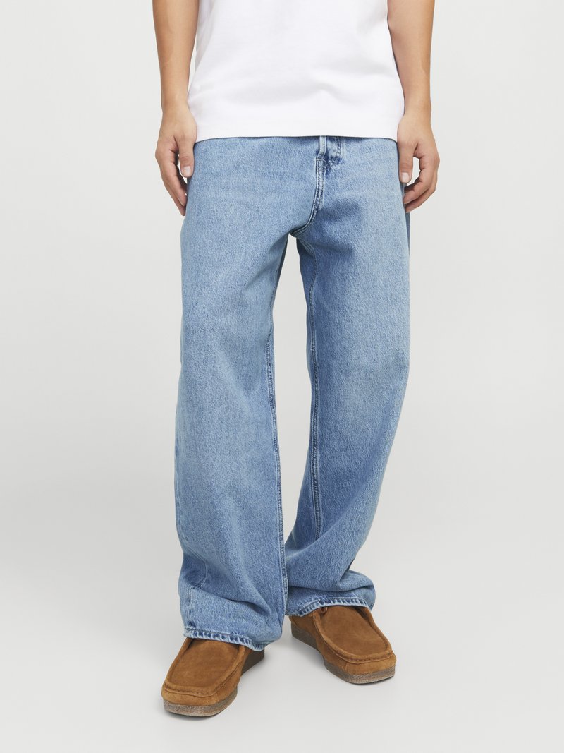 Vaqueros de pierna ancha en azul claro con un corte relajado, fabricados en mezclilla, con orillas deshilachadas, combinados con unos zapatos de ante marrón de tipo slip-on. Camiseta blanca sencilla.