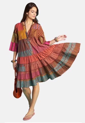 Woman wearing a colorful striped and patterned knee-length dress, holding the skirt out, with bracelets and a brown bag, standing barefoot.