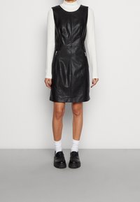 Schwarzes ledernes ärmelloses Kleid mit taillierter Silhouette, Nahtdetails an der Vorderseite und seitlichen Reißverschluss-Taschen, kombiniert mit einem weißen Langarmoberteil und schwarzen Schuhen.